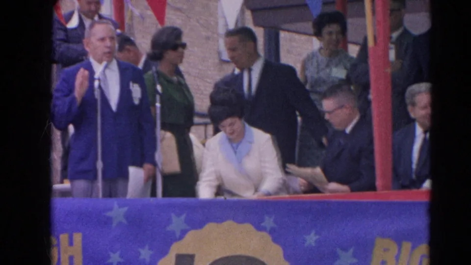 In 1964 Harvard, Illinois, a man in a blue suit stands at a silver microphone while formally dressed attendees sit and stand nearby.