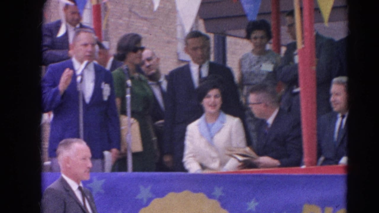 Cream-suited woman smiles near a microphone while men in suits and other attendees stand around her.