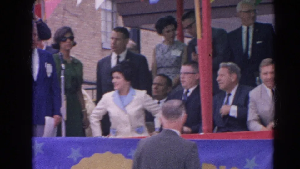 At the Harvard, Illinois public gathering, formal attendees on the platform shift positions while the speaker remains partly visible at left.