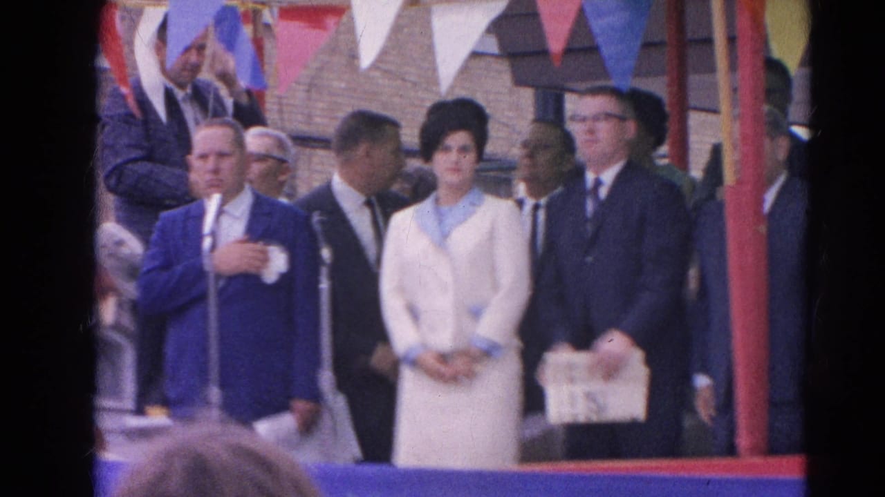 Dignitaries stand on a decorated outdoor stage with microphones and bunting.