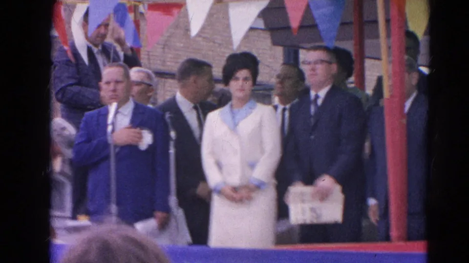 1964 Harvard, Illinois home-movie frame showing suited dignitaries and a woman in white on a bunting-decorated stage.