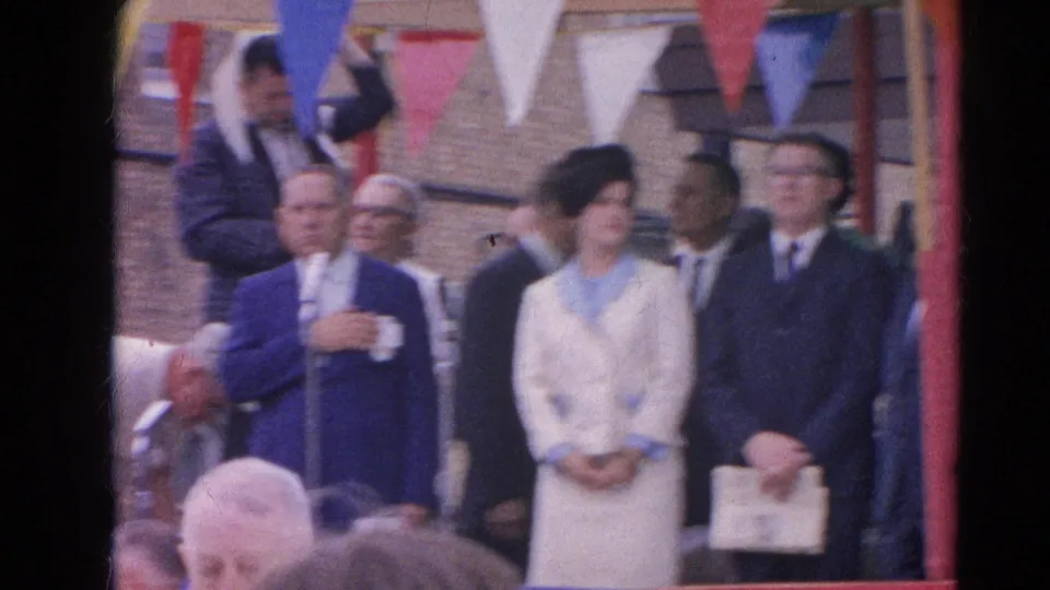 Home-movie frame from the 1964 Harvard, Illinois gathering, with dignitaries on stage and spectators partly blocking the lower view.