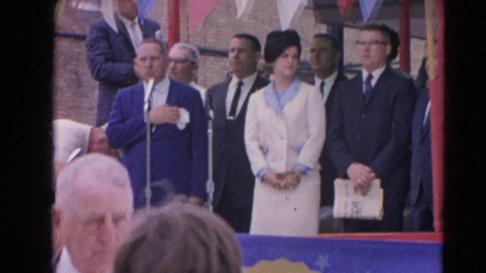 1964 Harvard, Illinois frame showing dignitaries lined up on an outdoor stage with microphones, bunting, and spectators below.