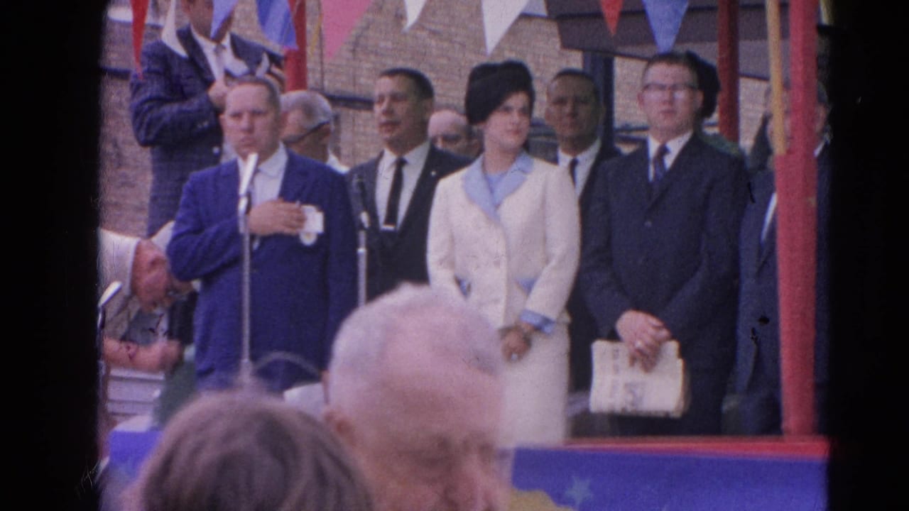 Man at microphones with formally dressed adults under patriotic bunting.