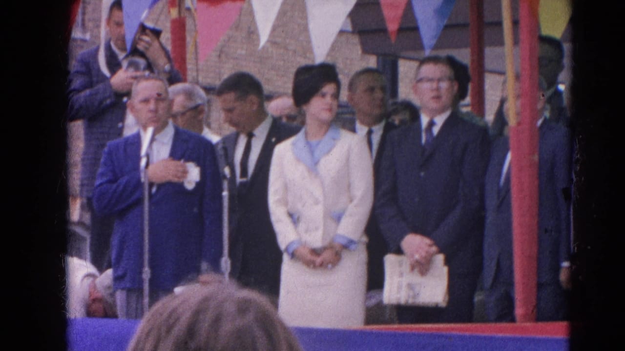 Ceremony platform with speaker, woman in white coat, and suited men.