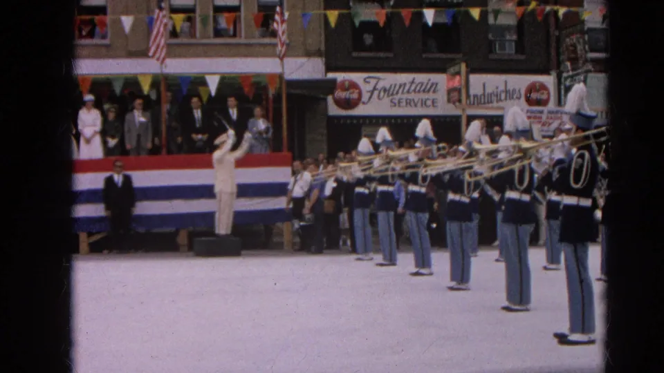 A marching band in blue and white uniforms performs on a Harvard, Illinois street in 1964, with spectators, flags, bunting, and storefront signs behind them.