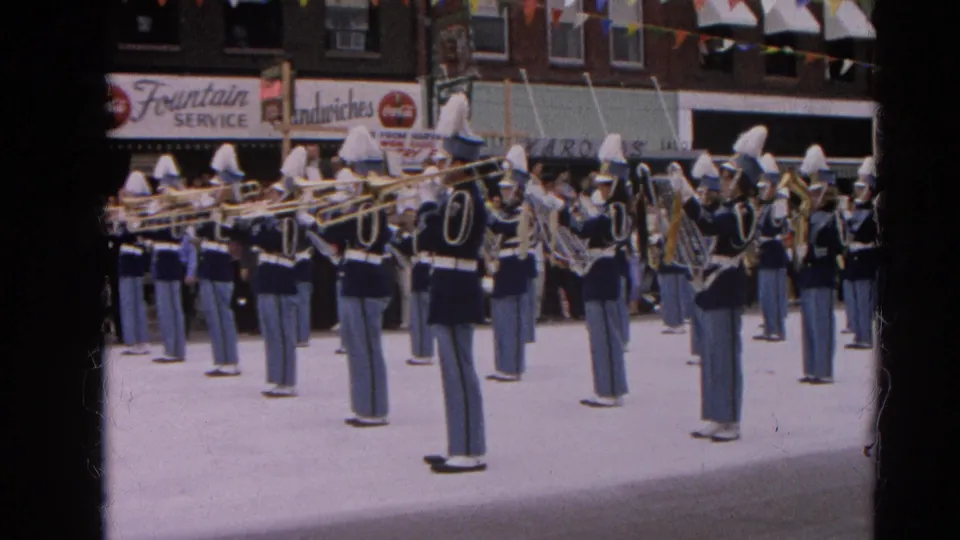1964 Harvard, Illinois parade view with marching band members in blue uniforms and white hats playing brass instruments near storefronts.