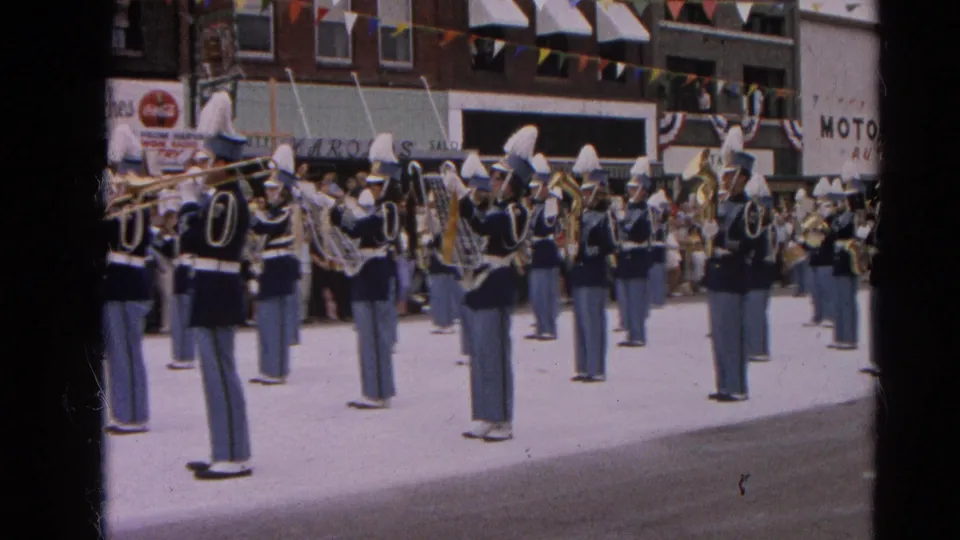 Close view of the marching band during the Harvard, Illinois parade, with brass players in blue uniforms and storefronts behind them.