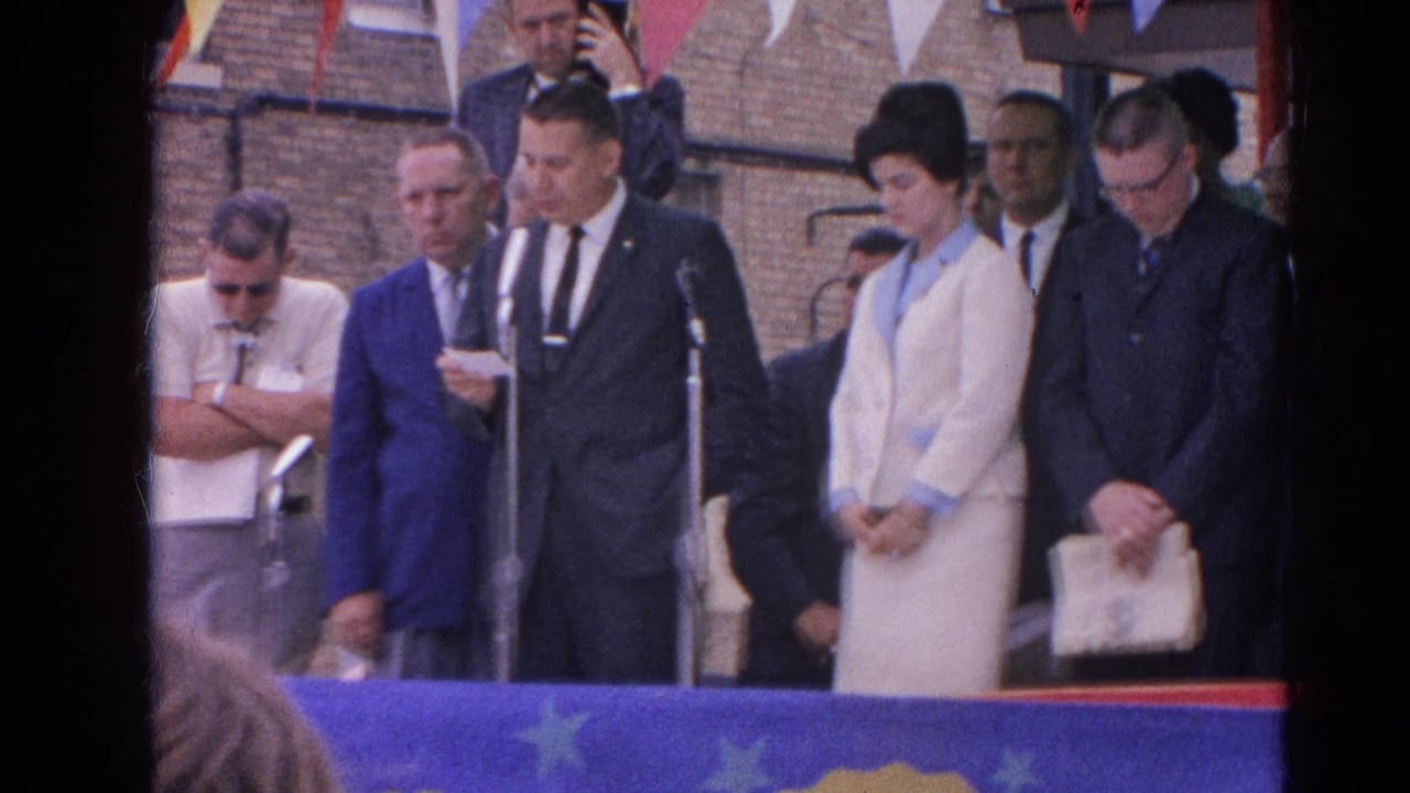 Suited adults stand at microphones during an outdoor ceremony behind a blue banner.