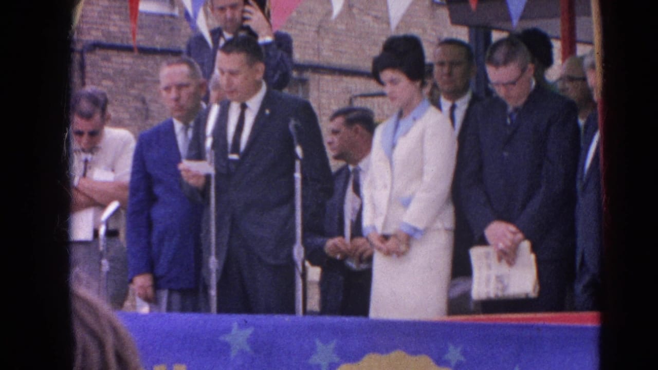 Adults stand behind a blue banner as a suited man reads near microphones.
