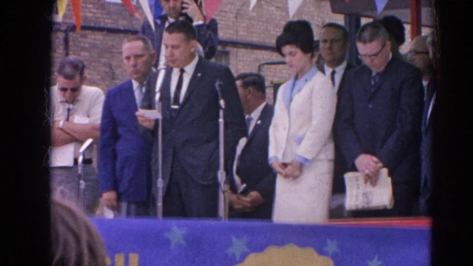 A Harvard, Illinois outdoor ceremony frame shows the speaker at microphones with a woman in a light suit and men in suits nearby.