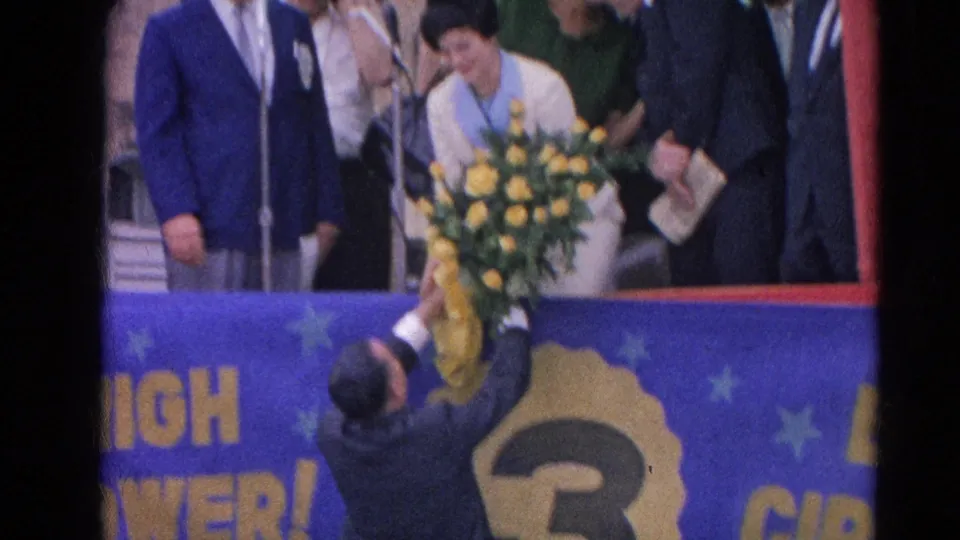At the 1964 Harvard, Illinois ceremony, a large yellow bouquet is handed up toward a woman and suited attendees near a blue banner.