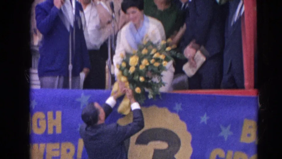 1964 Harvard, Illinois home-movie frame showing a woman on a decorated stage receiving yellow roses from a man below.