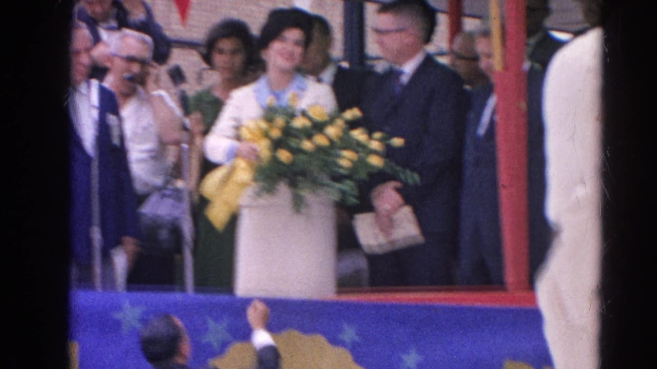 Woman in a light suit and dark hat holds a yellow bouquet on a decorated stage in Harvard, Illinois, 1964.