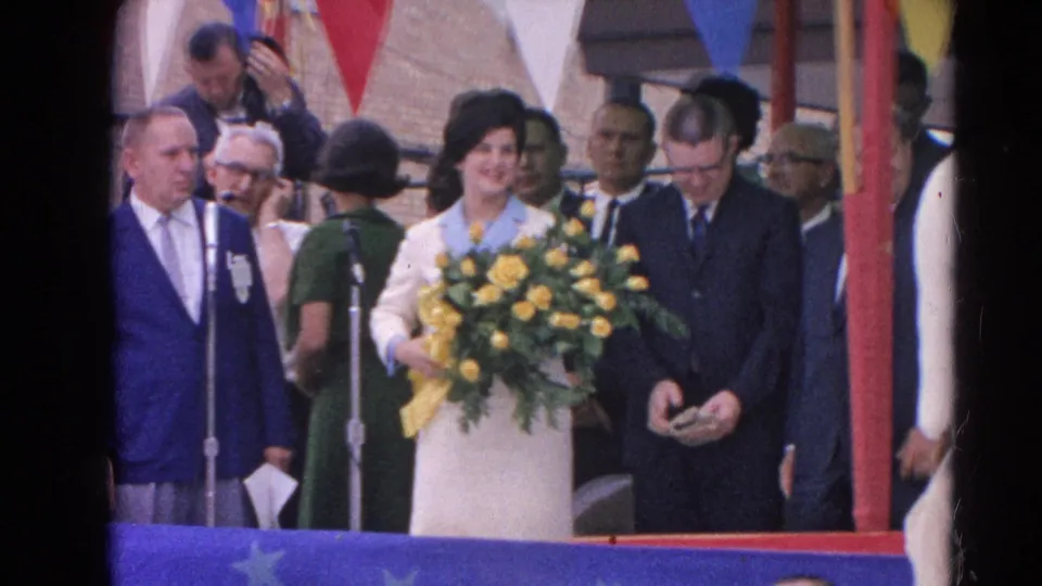Home-movie view of a smiling woman with yellow roses beside microphones at a 1964 public stage event in Harvard, Illinois.