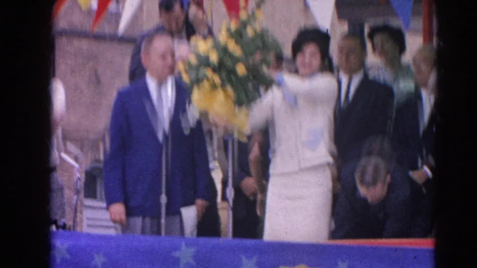 A woman lifts a large yellow bouquet on a decorated public stage as suited men stand nearby in 1964 Harvard, Illinois footage.