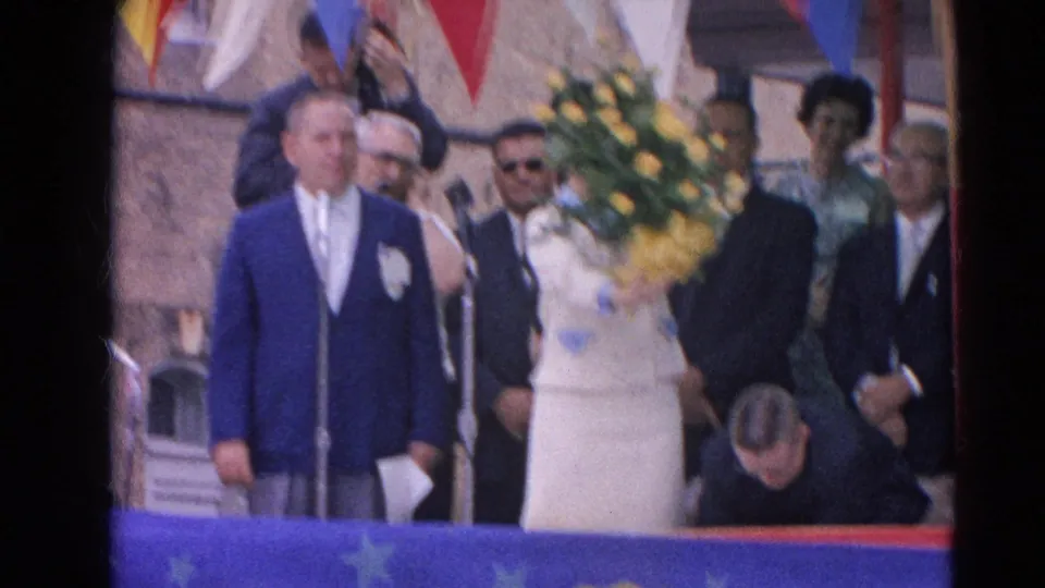 In Harvard, Illinois in 1964, a woman in white holds yellow flowers on a decorated platform beside suited men and a microphone.