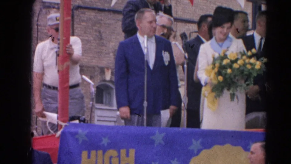 At the 1964 Harvard, Illinois outdoor ceremony, a woman with a bouquet stands beside suited men near a red pole and blue star banner.