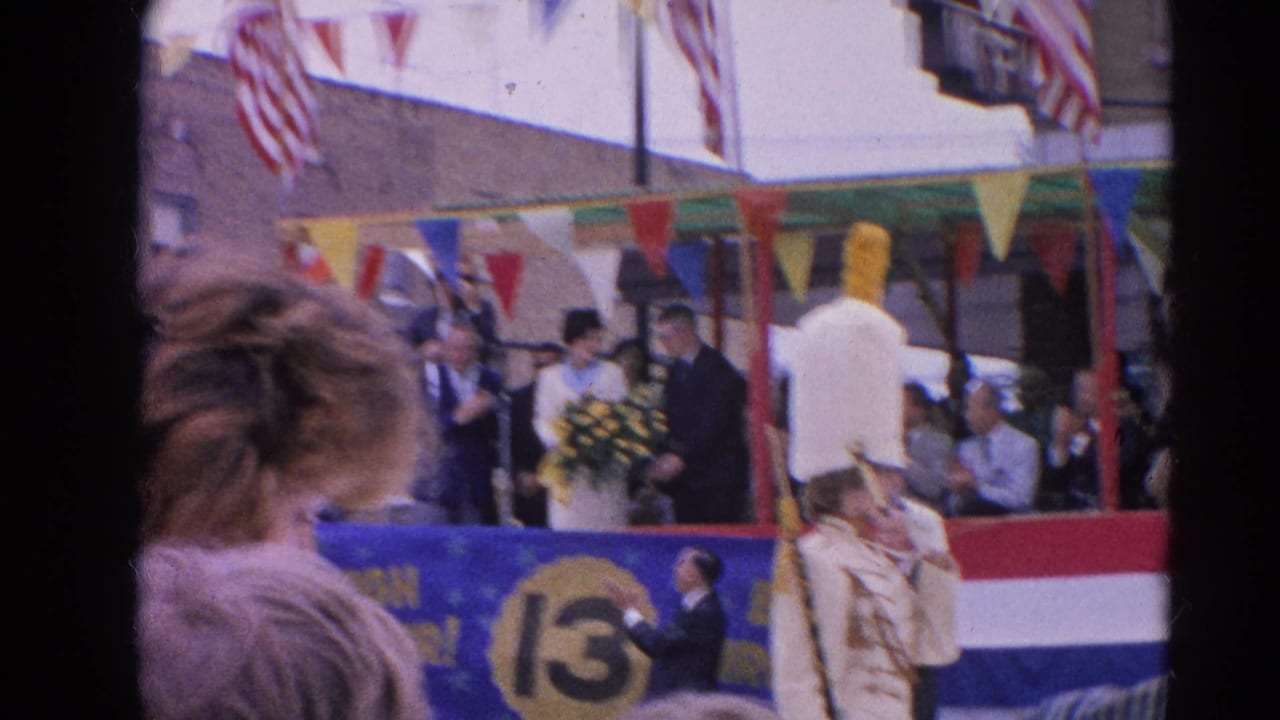 Crowd watches a decorated parade float with flags, bunting, and a white-uniformed marcher in front.