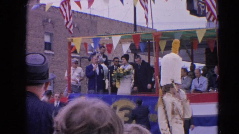 The Harvard, Illinois parade float is seen closer, with a woman holding a bouquet, suited participants, bunting, and spectators in the foreground.
