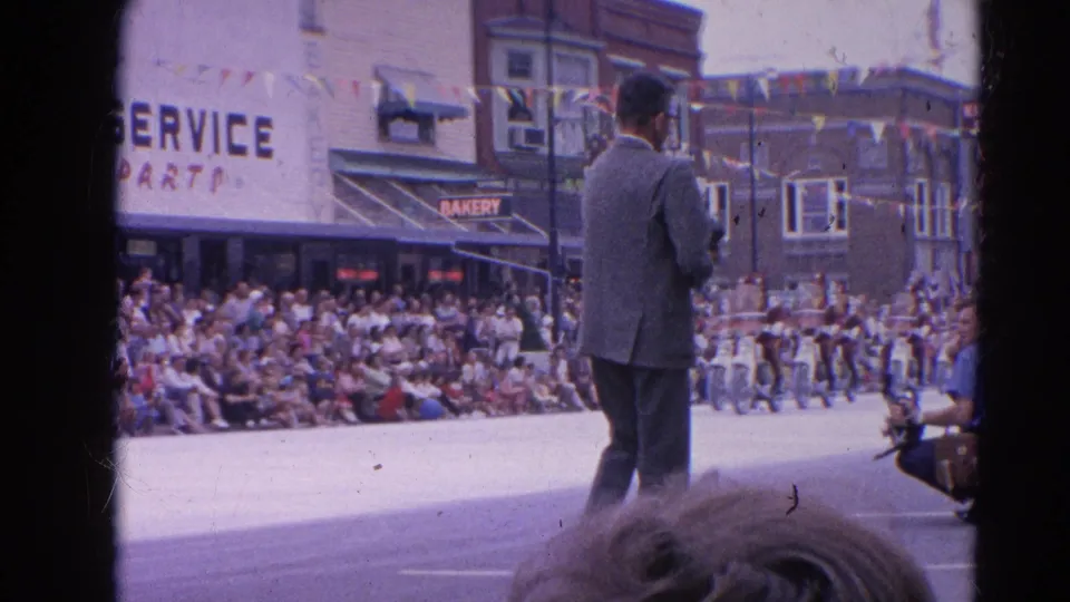 A man stands in the parade street in Harvard, Illinois, with seated spectators, storefront signage, bunting, and street decorations visible behind him.