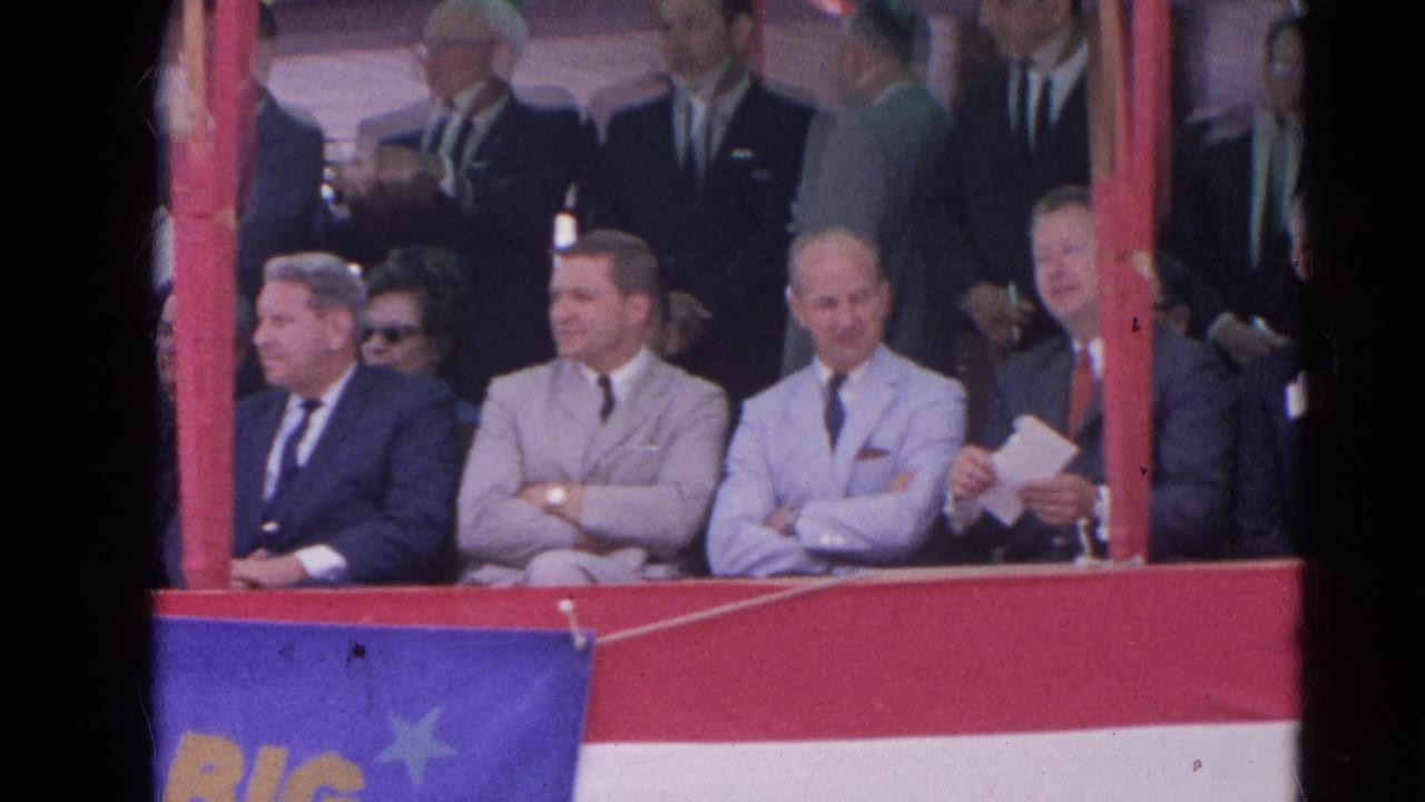 Men in suits sit in a decorated reviewing stand with red rails and blue fabric below.