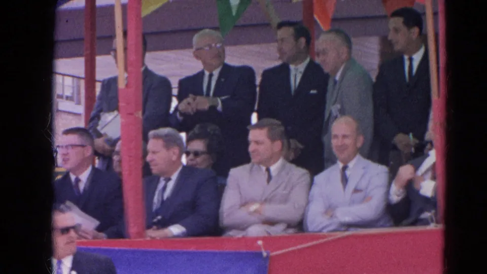 Seated men in 1960s suits watch from a decorated stand in Harvard, Illinois, with pennants overhead.