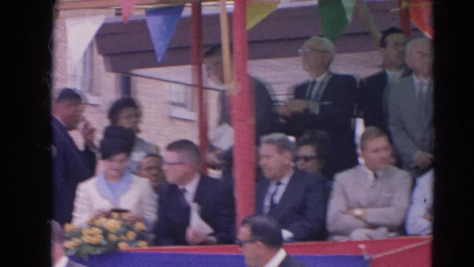 A woman holding yellow flowers sits among formally dressed spectators at the 1964 Harvard, Illinois festival stand.
