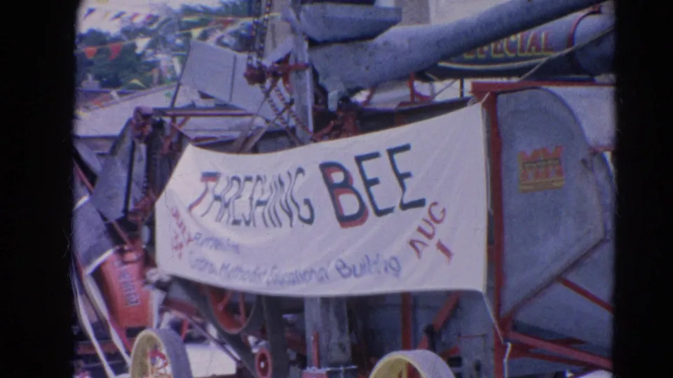 1964 Harvard, Illinois parade frame showing a Threshing Bee banner near equipment and crowd activity in soft 8mm color.