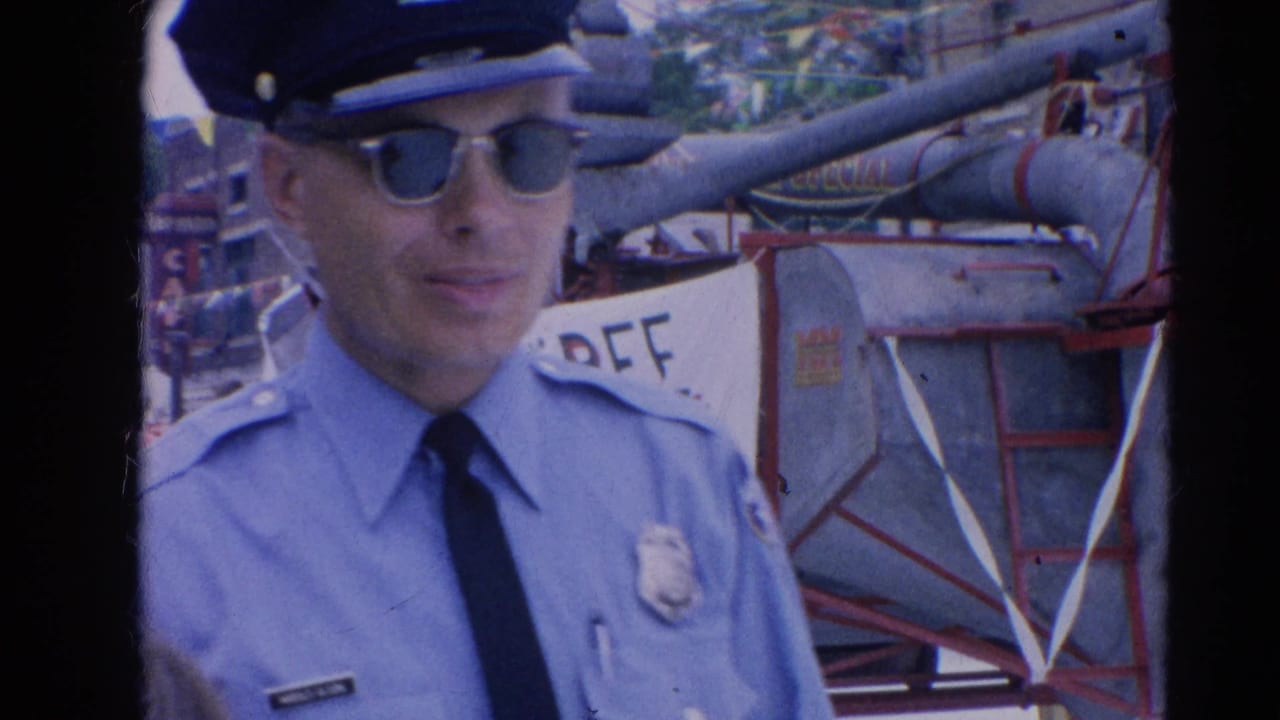 A uniformed police officer in sunglasses stands close to parade equipment and a Threshing Bee banner in 1964 Harvard, Illinois footage.