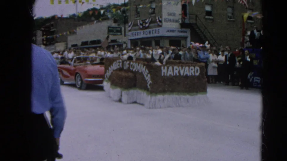 A Chamber of Commerce parade float marked Harvard passes crowds and storefronts in 1964 Harvard, Illinois home-movie footage.