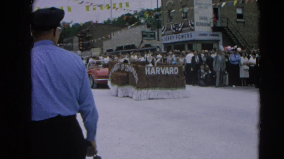 From behind a police officer, the frame shows the Harvard Chamber of Commerce float, a red Corvette, storefronts, and spectators.