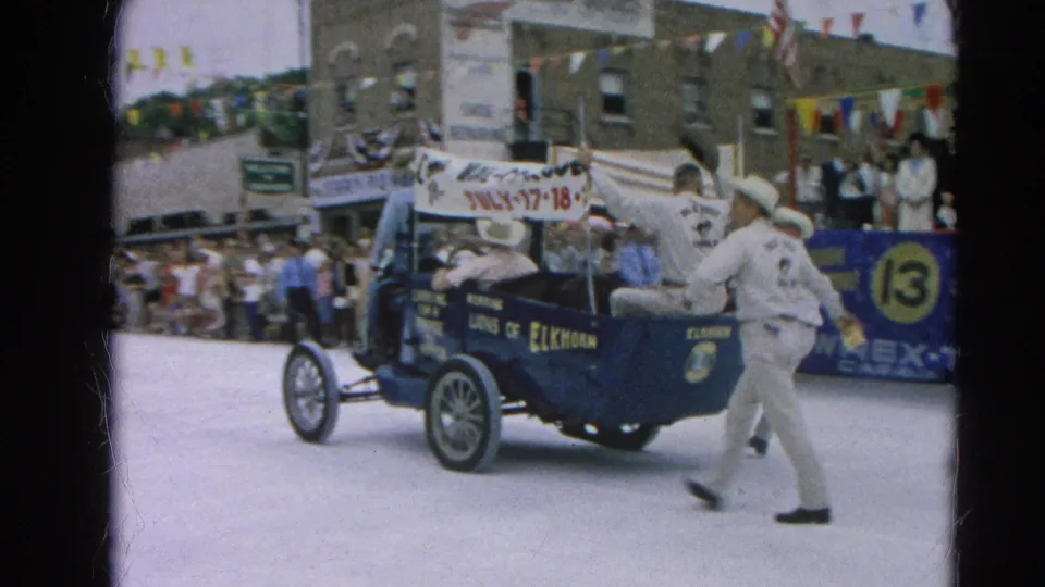 The Roaring Lions of Elkhorn car moves farther down the Harvard, Illinois parade route, with spectators, bunting, and a number 13 sign visible.