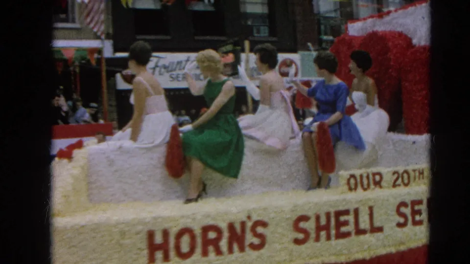 Close home-movie view of women in white, green, pink, and blue dresses seated on the Horn's Shell Service float in Harvard, Illinois.