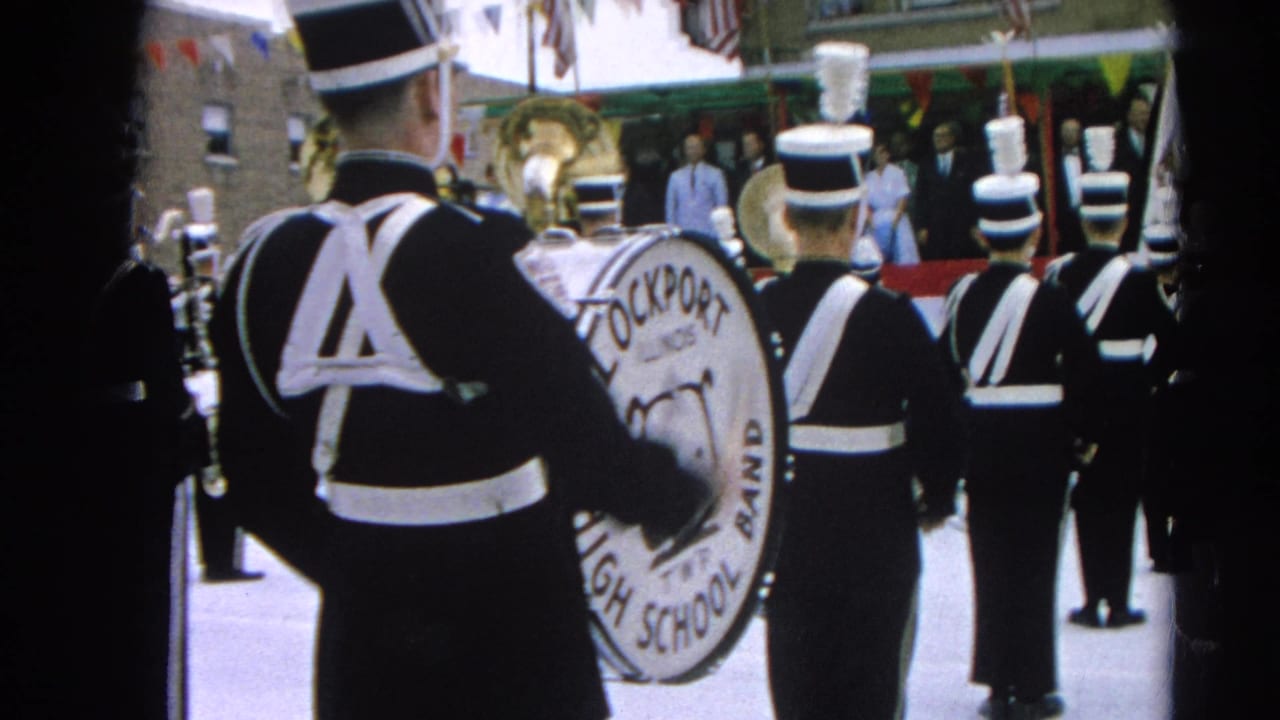 Uniformed marching band seen from behind with a bass drum and street decorations.