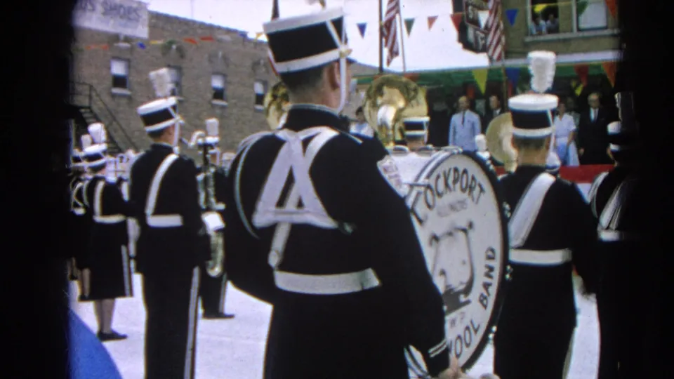 Final 1964 parade frame showing Lockport High School Band from behind on a decorated street in Harvard, Illinois.