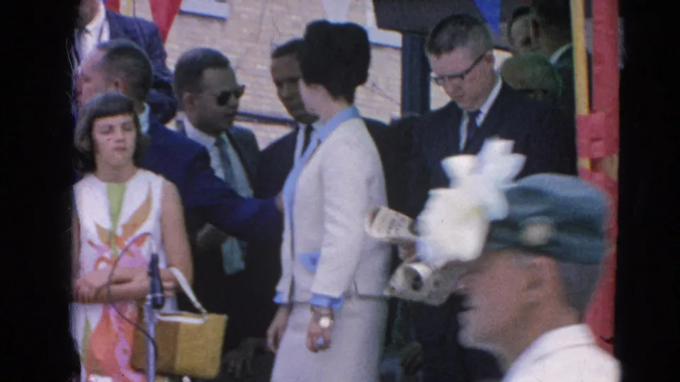 In Harvard, Illinois in 1964, a woman stands at a microphone as adults in 1960s clothing gather near the parade route.