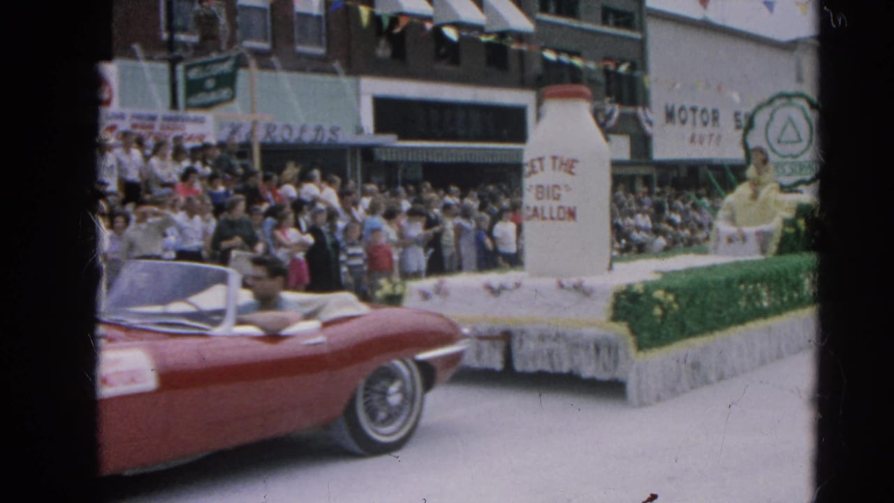 A red convertible moves past a Harvard, Illinois parade float with a giant milk bottle as spectators line the sidewalk in 1964.