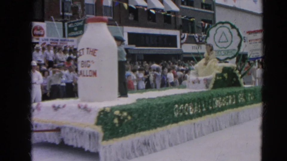 The dairy-themed parade float with a large milk bottle and sponsor lettering passes downtown spectators in Harvard, Illinois in 1964.