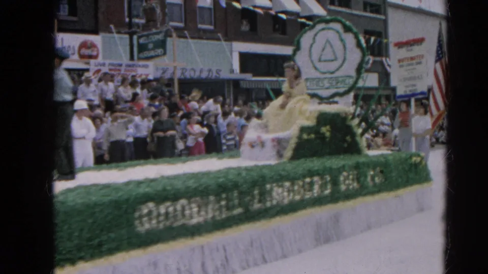 A parade float carrying a woman in a yellow dress passes spectators and storefronts in Harvard, Illinois during the 1964 street parade.
