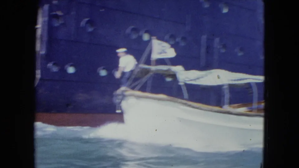 The launch moves partly out of frame beside the dark blue ship hull, with a sailor forward and water spray along the boat's side.