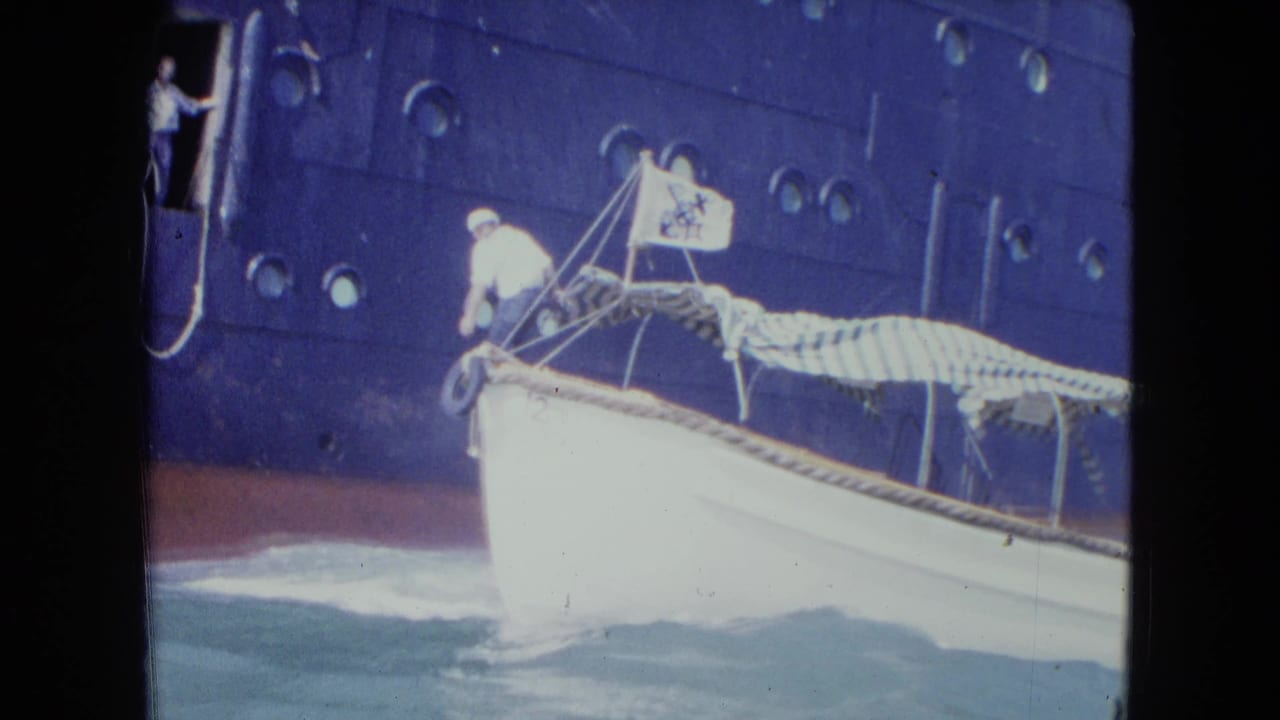 1964 Kingston, Jamaica home-movie frame showing a white launch approaching a dark blue ocean liner with crew and doorway observers visible.