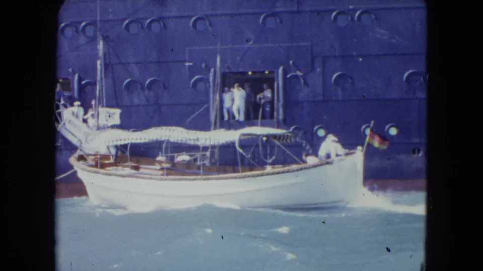 1964 Kingston harbor frame showing a small white boat with striped canopy and stern flag passing beside a dark ocean liner.