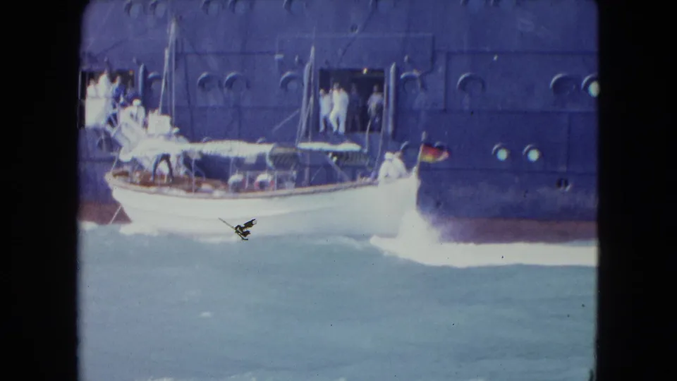 In 1964 Kingston, Jamaica, a small white motorboat passes alongside a dark blue ocean liner as people stand near an open side doorway.