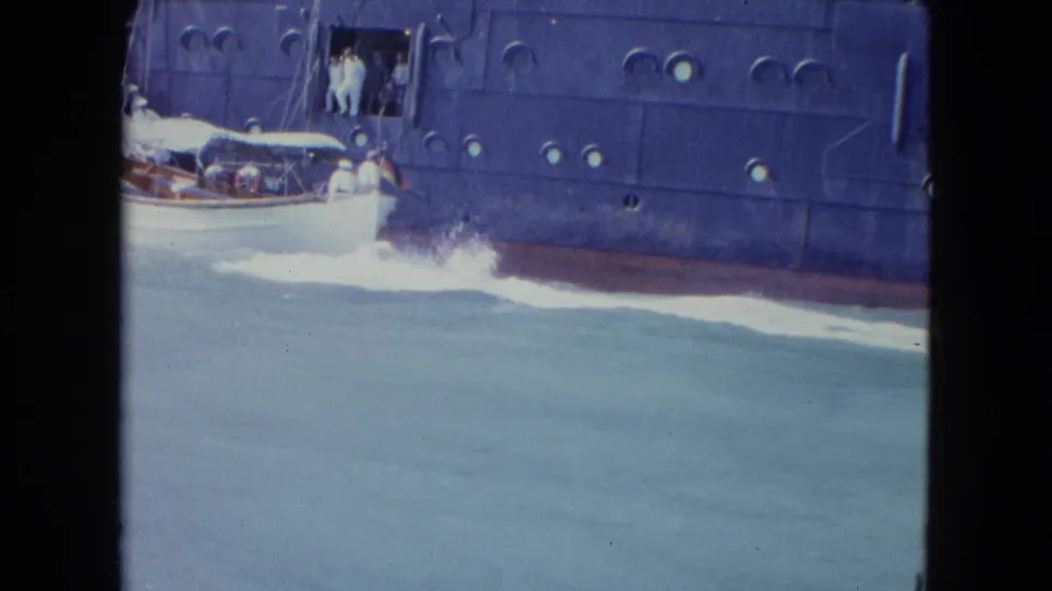 The motorboat continues past the liner in the Kingston harbor footage, partly cropped at the left edge while white wake spreads behind it.