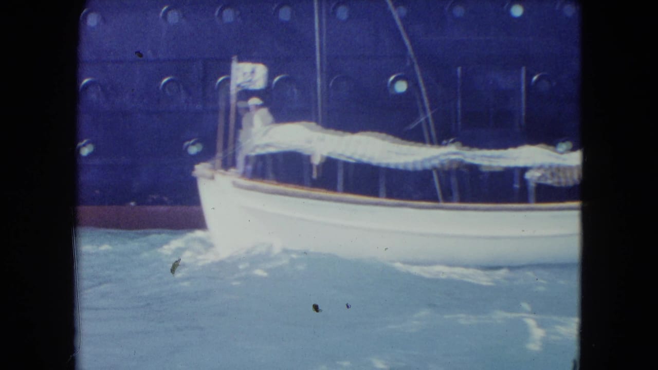 White boat side-on in rough water with striped awning and large ship behind it.