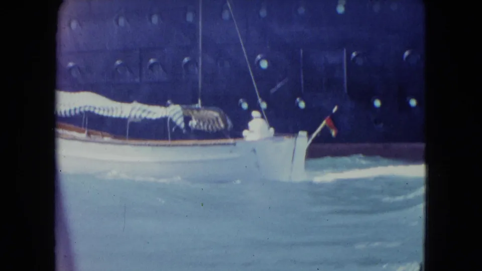 The stern end of the white boat rolls through rough Kingston harbor water near a dark ship hull, with a small flag visible at the bow area.