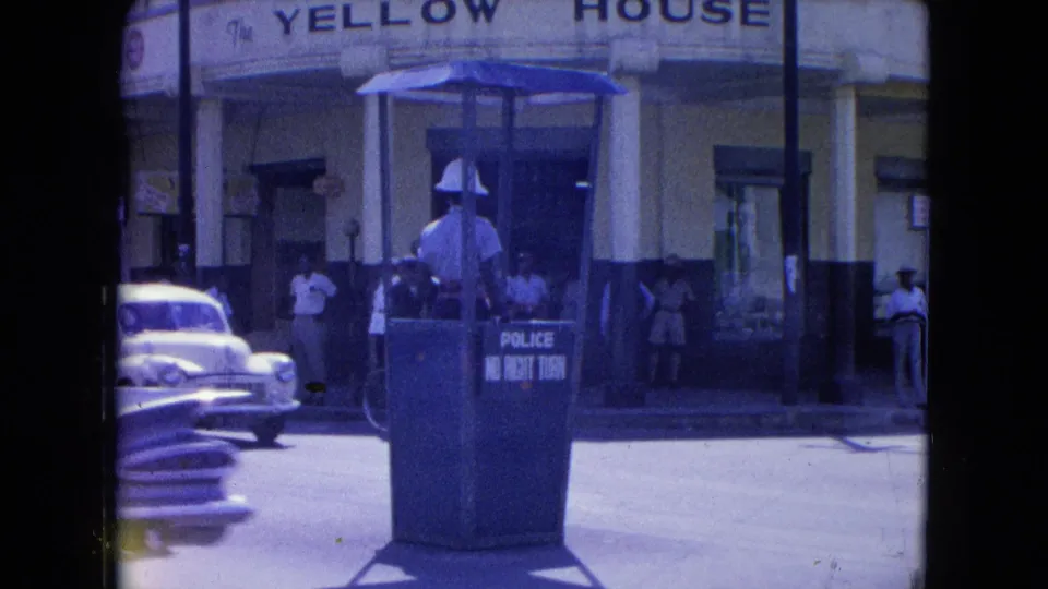 1964 Kingston street scene with a traffic officer booth, passing mid-century cars, pedestrians, and The Yellow House sign.