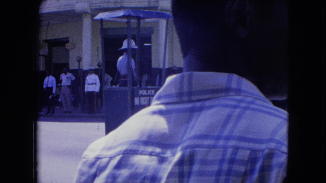 In Kingston, Jamaica, a foreground man in a plaid shirt partly obscures a white-uniformed officer standing in a raised traffic booth.