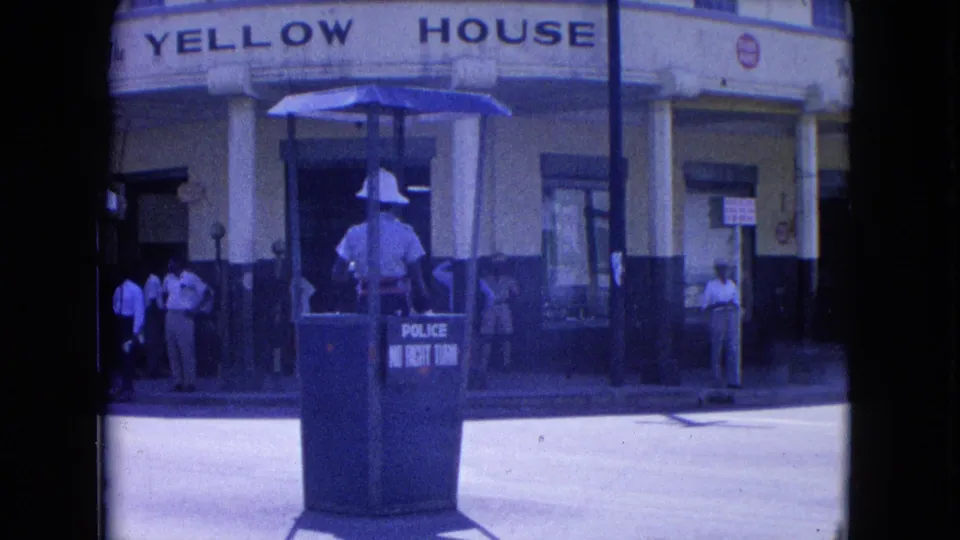 In Kingston, Jamaica, a police officer in white uniform stands in a blue booth marked for traffic control outside the Yellow House.
